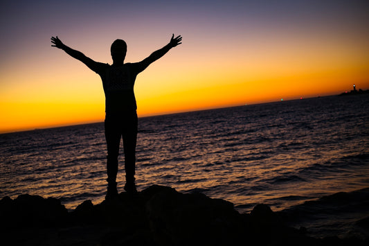 silhouette of woman standing on rock formation near body of water during sunset