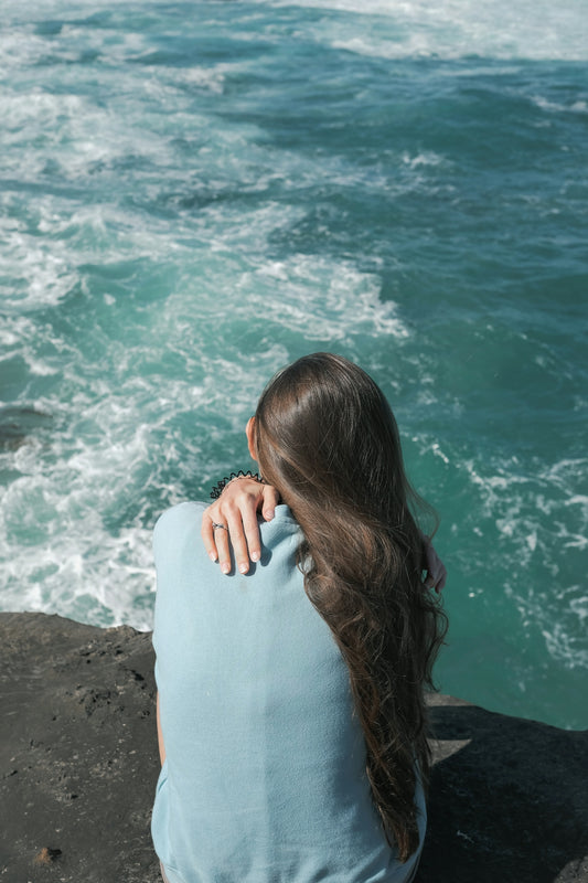 woman in white long sleeve shirt sitting on rock near body of water during daytime