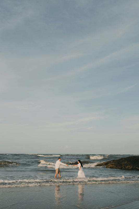 A couple walks along a beach, holding hands.
