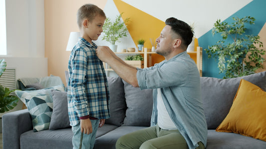 Father buttons son's plaid shirt while sitting on couch.