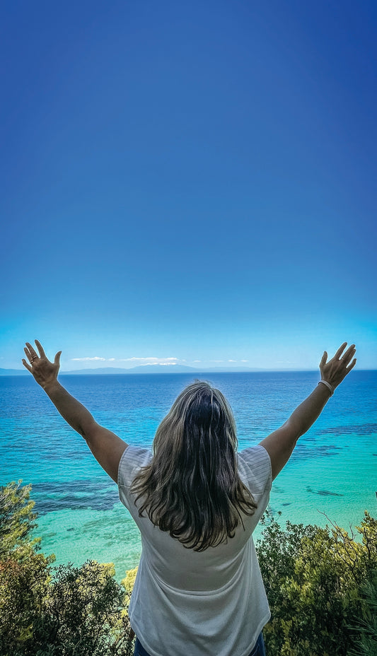 Woman with arms raised overlooking a vibrant blue ocean.
